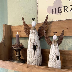 May include: Two decorative angel figurines crafted from white distressed wood with rusted metal wings and wire halos. Each angel features a small heart-shaped cutout in the center of its body. A dark gray candle sits in a brass-colored metal holder.
