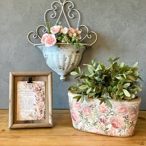 Peut inclure: Composition décorative avec une jardinière murale blanche vieillie avec des roses roses et de la verdure, une image encadrée en bois avec une impression florale et une jardinière à motifs floraux avec de la verdure.