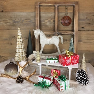 May include: A white rocking horse, a wooden window frame, a metallic ornament, decorative trees, a small sled, and wrapped gifts. The scene includes a gold antler, a pinecone, and a mercury glass sphere, all arranged on a white faux fur surface.