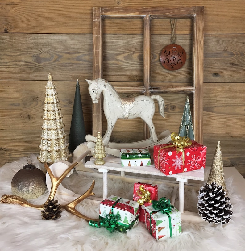 May include: A white rocking horse, a wooden window frame, a metallic ornament, decorative trees, a small sled, and wrapped gifts. The scene includes a gold antler, a pinecone, and a mercury glass sphere, all arranged on a white faux fur surface.