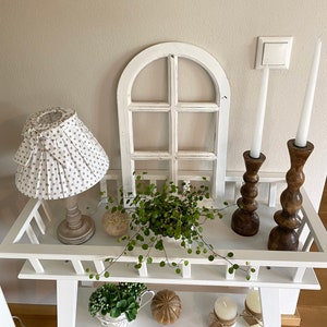 May include: A white wooden console table featuring a lamp with a beige polka dot shade, a white arched window frame, a green plant, and two dark brown wooden candle holders with white taper candles. The lower shelf holds a plant and two candles.