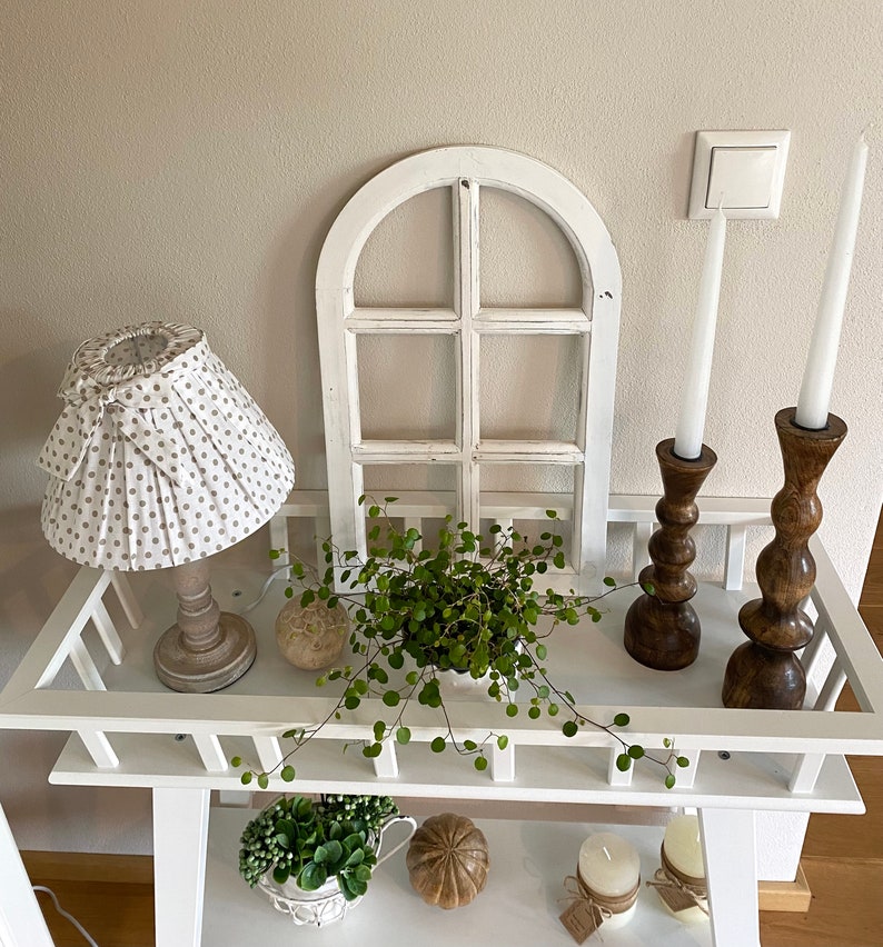 May include: A white wooden console table featuring a lamp with a beige polka dot shade, a white arched window frame, a green plant, and two dark brown wooden candle holders with white taper candles. The lower shelf holds a plant and two candles.