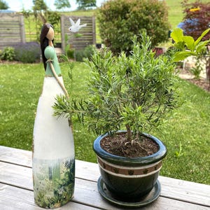 Könnte beinhalten: Eine große, schlanke Dekofigur einer Frau mit dunklem Haar, grünem Oberteil und weißem Rock mit botanischem Aufdruck, die neben einer kleinen grünen Topfpflanze in einem dunkelgrünen Keramiktopf mit passendem Untersetzer steht.