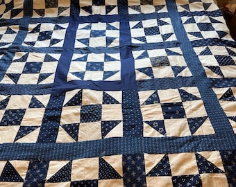 May include: A handmade quilt featuring a patchwork design in shades of white and navy blue. The quilt showcases a repeating pattern of squares and triangles, with a navy blue border. The quilt is spread out on a bed.