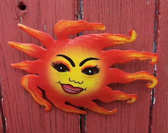 May include: A red, yellow and orange metal sun with a smiling face. The sun has a yellow face with black eyes, a black nose and a red mouth.