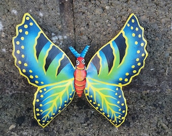 May include: A vibrant butterfly ornament, featuring blue, green, yellow, black, and red hues. The butterfly's wings display a detailed pattern and are set against a textured grey backdrop. The wings are decorated with yellow spots.
