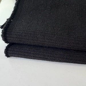 May include: Folded black knitted fabric, possibly a scarf or wrap, with a ribbed texture. The fabric is a solid, dark color, and appears to be made of a soft material. The edges are slightly frayed.