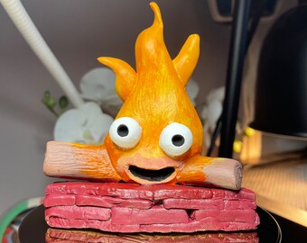 May include: A hand-painted clay figurine of a smiling orange fire with white eyes, sitting on a red brick base. The fire is on top of two brown logs.