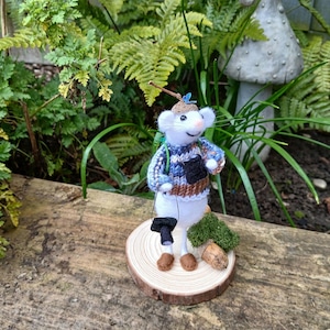 May include: A felt mouse figure wearing a blue, brown, and white knitted sweater, backpack, and hat. The mouse holds a black bucket and stands on a wooden slice with moss and an acorn. The background includes greenery and a mushroom statue.