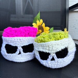 May include: Two crochet skull-shaped containers, one with a pink top and the other with a green top. The skulls are white with black eye sockets. A yellow sunflower and green leaves are in the background.