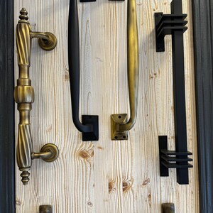 May include: Five different door handles displayed on a white wood panel. The handles are made of metal and have a variety of styles and finishes, including black, brass, and gold.