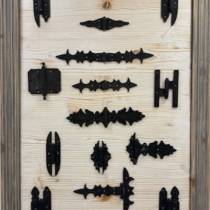 May include: A collection of 11 black metal hinges with various decorative designs, displayed on a light wood background.