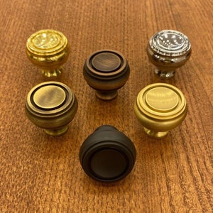 Op de afbeelding: Zes verschillende kastknoppen in verschillende afwerkingen, waaronder messing, brons en zwart. De knoppen zijn rond met een verhoogd midden en een decoratieve rand.
