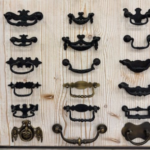 May include: A collection of 14 different black and brass drawer pulls and handles. The pulls and handles are displayed on a white wooden surface.