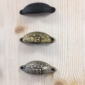 May include: Five different metal cabinet pulls with a decorative buckle design. The pulls are arranged vertically in a row from top to bottom. The pulls are in the following finishes: copper, black, gold, silver, and brass.