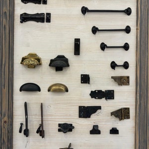 May include: A display of various black and brass door hardware, including hinges, latches, and handles. The hardware is arranged on a white wooden board with a dark wood frame.