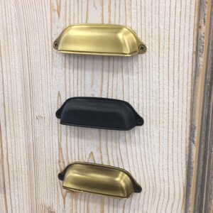 May include: Five different cabinet pulls in various finishes. The pulls are all the same shape, a slightly curved rectangle with rounded edges. The finishes are copper, gold, black, brass, and nickel.