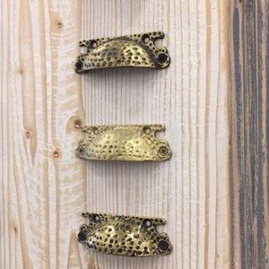 May include: Five different metal cabinet pulls with a hammered finish. The pulls are arranged vertically on a wooden surface. The pulls are black, gold, brass, and copper.