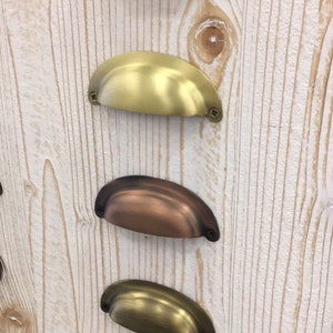 May include: Five different cabinet pulls in various finishes. The pulls are all the same shape, a rounded rectangle with a slight curve in the center. The finishes are black, gold, copper, brass, and nickel.