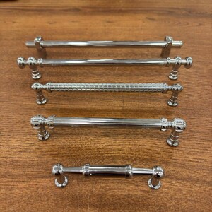 May include: Five different styles of silver cabinet pulls, all with rounded ends. The pulls are arranged in a vertical line on a wooden surface.