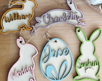 May include: Five wooden Easter decorations with names written on them. The decorations are shaped like bunnies and eggs. The names are William, Charlotte, Jace, Maddy, and Sophia. The decorations are painted in various pastel colours.