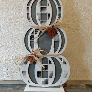 Może przedstawiać: Stos trzech białych drewnianych dyni w szachownicę w kolorze szarym i białym. Każda dynia ma mały brązowy liść przymocowany do góry. Dynie stoją na białej podstawie.