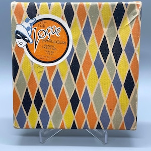 May include: A square box featuring a repeating diamond pattern in orange, yellow, blue, and grey. A circular label on the top left displays a harlequin illustration and text reading IT'S THE VOGUE HARLEQUIN PENCIL TABLE NO. TABLES 1 TO 6 11577 THE BUZZA CO MPLS. U.S.A.