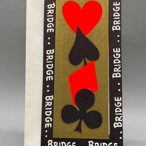 May include: A greeting card with a gold background featuring playing card suits: a red heart, spade, diamond, and club. The word "BRIDGE" is printed in white around the edges of the card. The card is propped up on a clear stand.