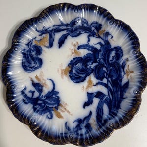 May include: A round ceramic plate with a scalloped edge, featuring a white base decorated with deep blue floral patterns and gold accents. The rim is finished with a dark blue wash and gold detailing, creating a textured appearance.