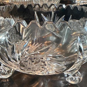 May include: A clear, cut-glass bowl with a scalloped rim and three decorative feet. The bowl features intricate starburst and leaf patterns, reflecting light. A decorative piece for serving or display.