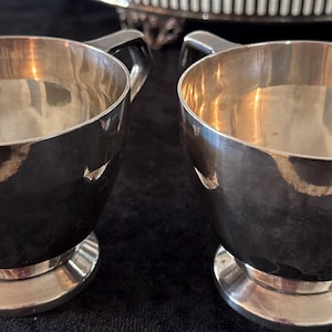 May include: Two polished silver-toned metal cups with a smooth, reflective finish. Each cup features a wide, open bowl shape that tapers down to a circular pedestal base and includes a single curved handle attached to the side of the vessel.