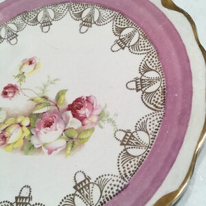May include: A white porcelain plate with a pink and gold rim. The plate features a floral design with pink and yellow roses and green leaves. The rim is decorated with a gold lace pattern.