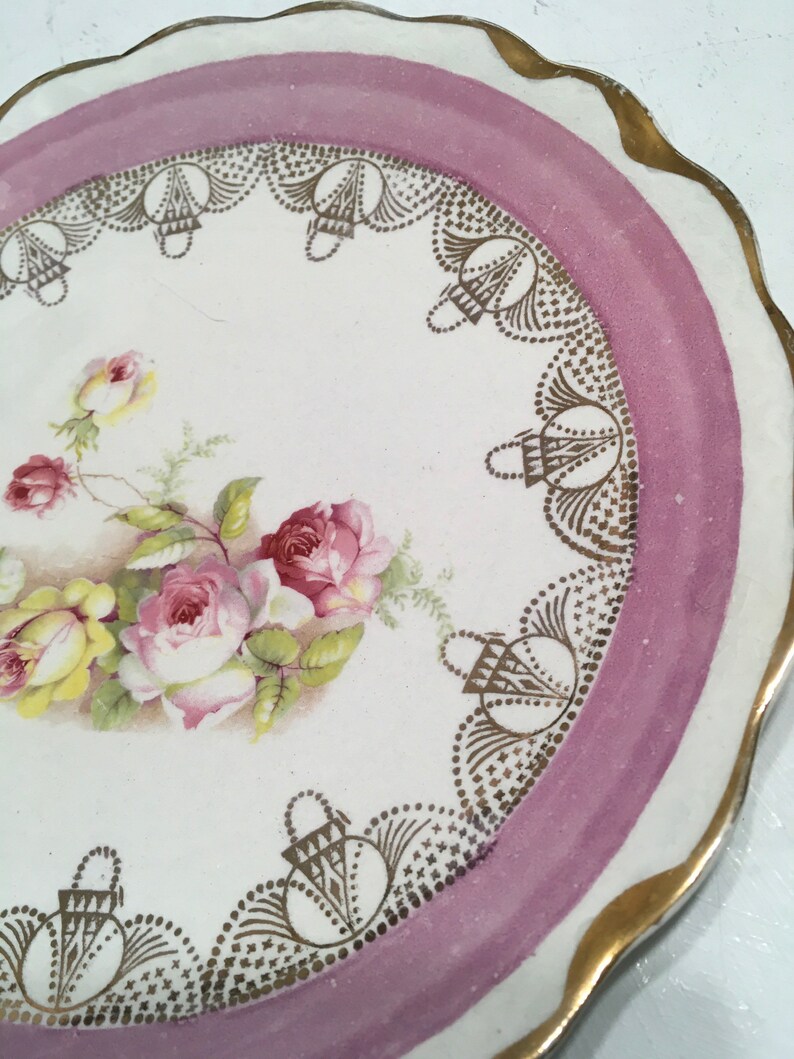 May include: A white porcelain plate with a pink and gold rim. The plate features a floral design with pink and yellow roses and green leaves. The rim is decorated with a gold lace pattern.