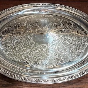 May include: An oval silver-colored serving tray featuring ornate embossed scrollwork and shell motifs across the surface. The tray includes two decorative handles on the sides and a smooth, polished central area for serving items.
