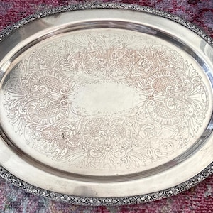 May include: An oval silver-toned serving tray featuring ornate embossed floral and scrollwork patterns across the center surface. The tray includes two decorative handles on the sides and a detailed textured border along the outer rim.