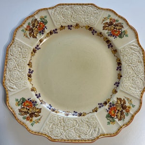 May include: A cream-colored ceramic plate with a scalloped edge and a floral design. The plate features a border of purple berries, green leaves, and orange flowers.