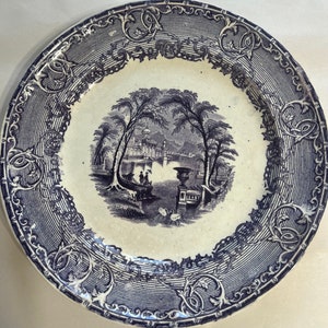 May include: A white ceramic plate with a blue and white design. The plate features a scene of a garden with trees and a building in the background. The plate is decorated with a blue and white floral border.