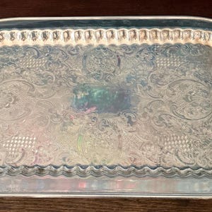 May include: A rectangular silver-toned serving tray featuring ornate embossed floral and scrollwork patterns across the surface. The tray has a scalloped edge and two decorative, curved metal handles attached to the short sides.