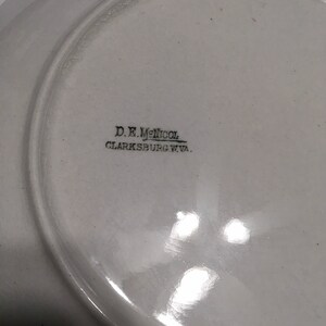 May include: White ceramic plate with black text on the bottom that reads "D.E. McNicol Clarksburg W.Va."