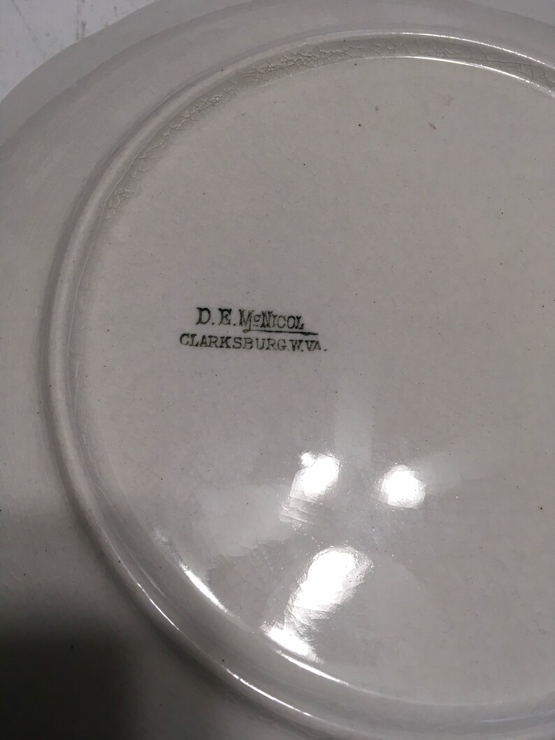 May include: White ceramic plate with black text on the bottom that reads "D.E. McNicol Clarksburg W.Va."