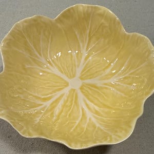 May include: A pale yellow ceramic bowl shaped like a cabbage leaf. The bowl features a glossy finish with embossed, raised white veins radiating from the center toward the scalloped, wavy edges, mimicking the natural texture of a leafy vegetable.