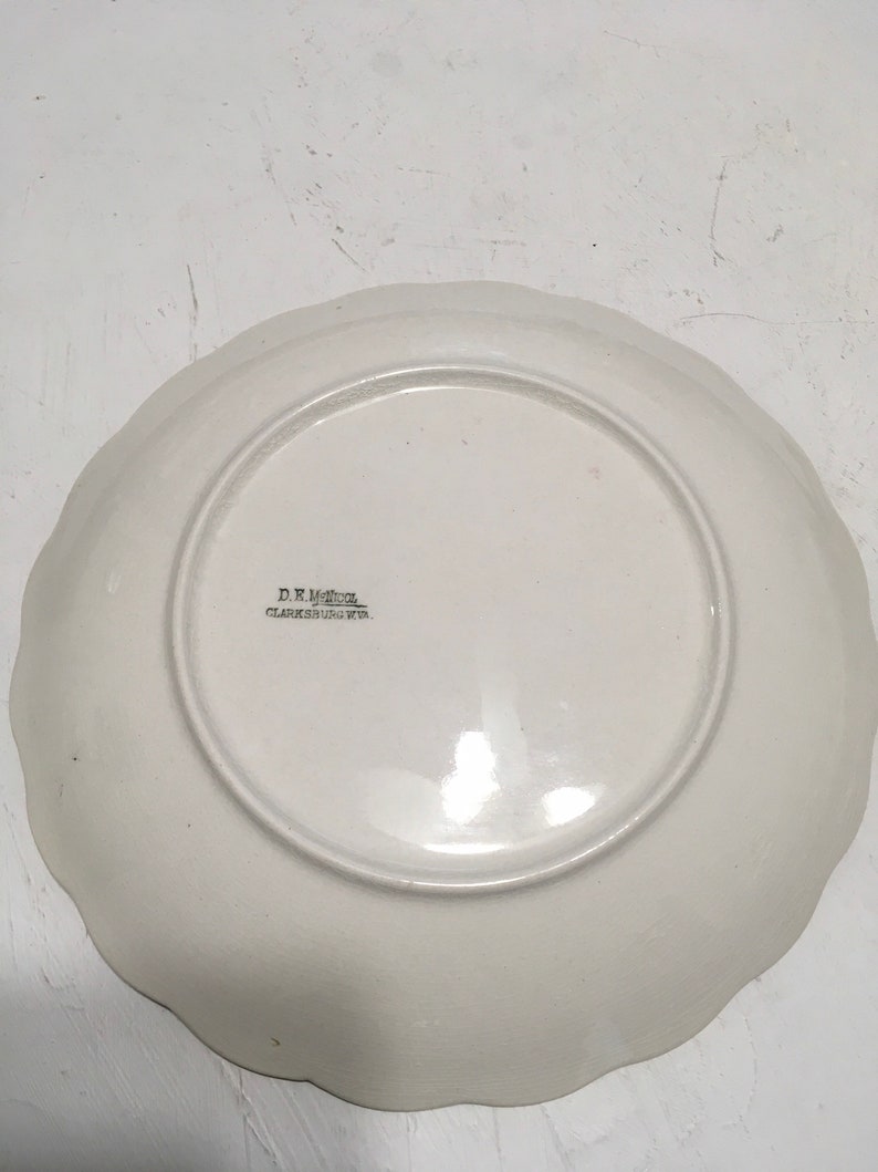 May include: White ceramic plate with a scalloped edge. The plate has a floral design and the text "D.R. Miller, Clarksburg, W.Va." printed on the bottom.