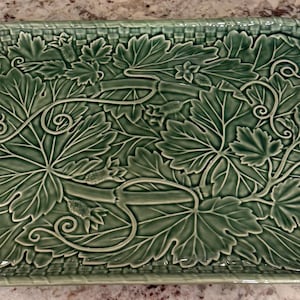 May include: A rectangular ceramic serving tray in a sage green glaze featuring a raised, embossed pattern of grape leaves, curling vines, and small clusters of grapes. The border of the tray has a textured, woven basket-weave design.