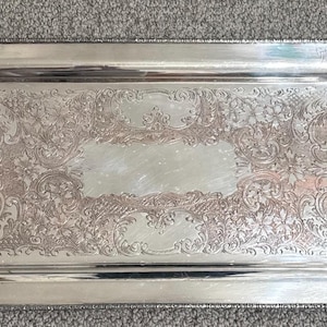 May include: A rectangular silver-toned serving tray featuring ornate floral and scrollwork engravings across the center surface. The tray includes two integrated handles on the sides and a decorative beaded edge along the outer rim.