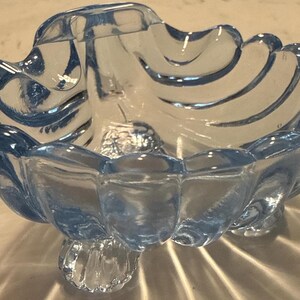 May include: A light blue, scalloped-edge glass dish with three decorative feet. The dish has a central, raised design and a wavy, shell-like pattern around the rim. The glass is transparent, allowing light to pass through.