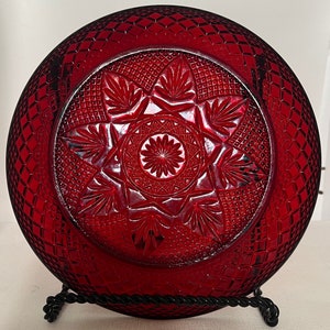 May include: A deep red glass plate with a geometric pattern. The plate has a raised center with a starburst design.