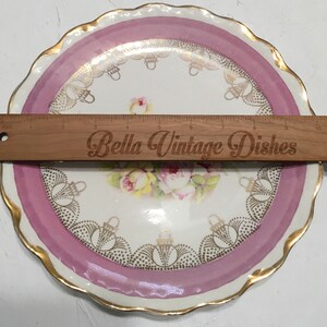 May include: A vintage porcelain plate with a pink rim and gold accents. The center features a floral design with roses and a gold basket pattern. The plate is 9 inches in diameter.
