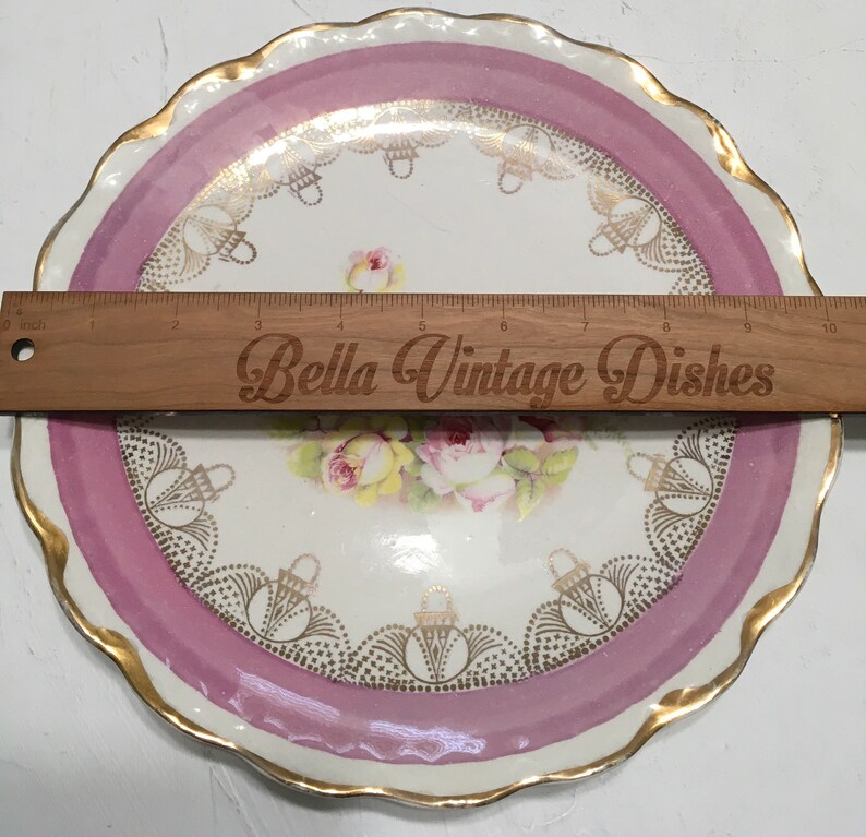 May include: A vintage porcelain plate with a pink rim and gold accents. The center features a floral design with roses and a gold basket pattern. The plate is 9 inches in diameter.