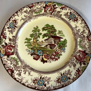 May include: A vintage ceramic plate with a brown and cream floral pattern. The center of the plate features a cottage scene with a brown roof and green trees.
