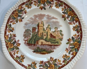 May include: A white ceramic plate with a brown, green, and yellow floral border. The center of the plate features a hand-painted image of a stone castle with a river in front of it.